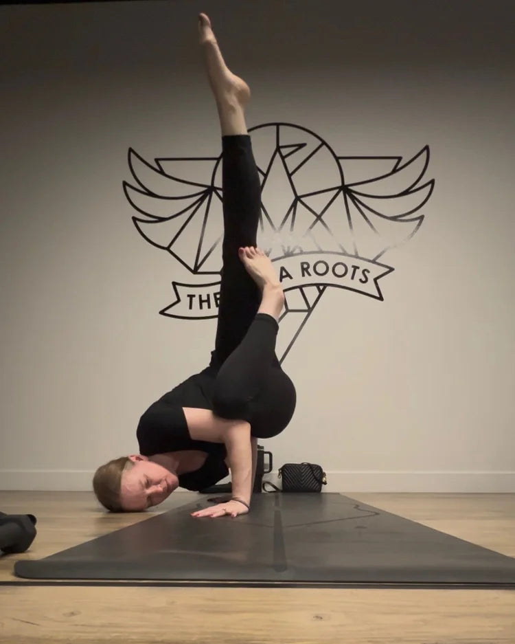 Jen Montgomery in fallen angel yoga pose on a black yoga mat in a studio, wearing a black short-sleeve top and black leggings. The background features a light gray wall with a black winged logo and the text 'THE YOGA ROOTS'.
