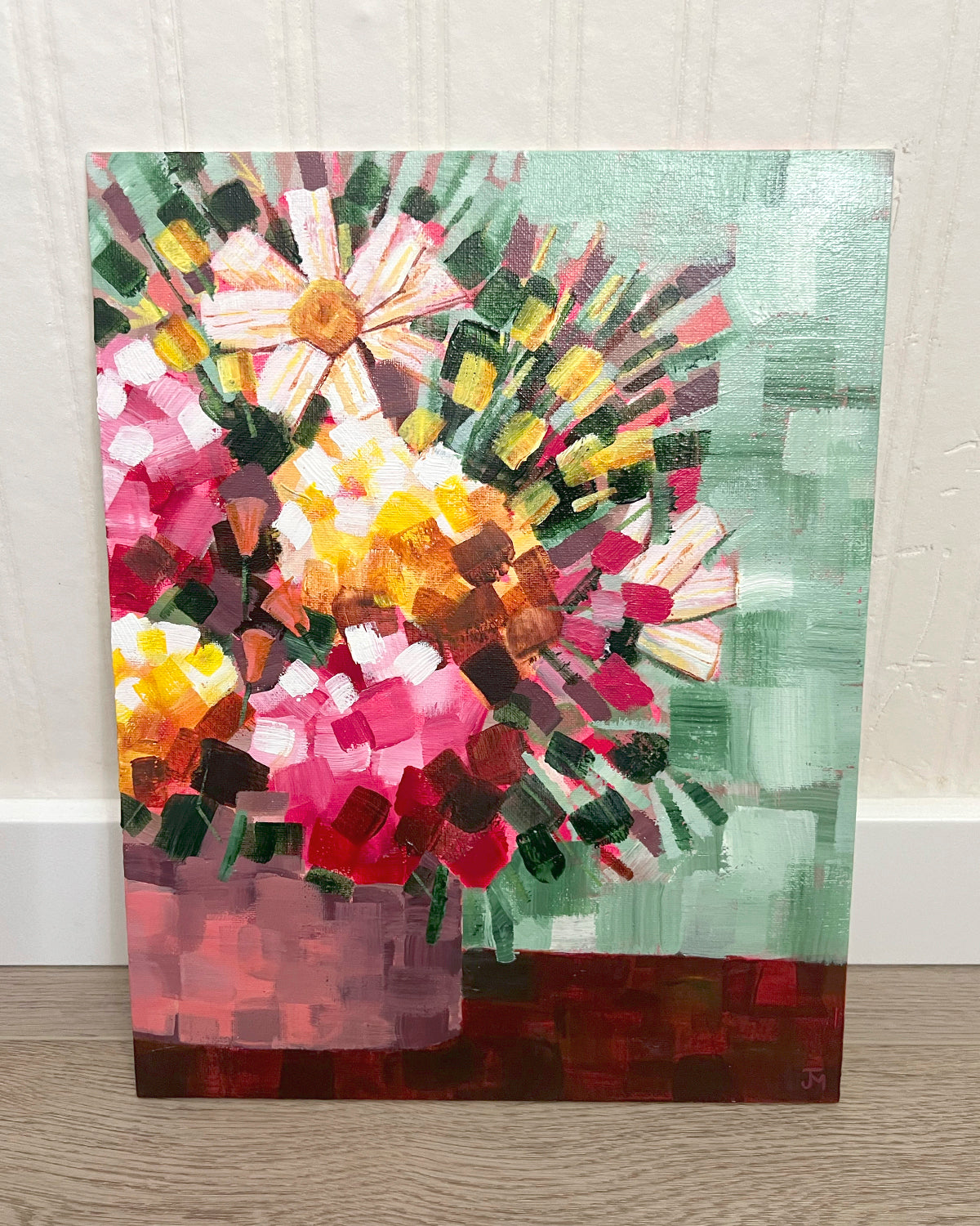 A close-up of an abstract floral painting by Jen Montgomery on canvas, showing textured brushstrokes in pink, red, yellow, green, and brown, leaning against a white wall on a light wood floor.