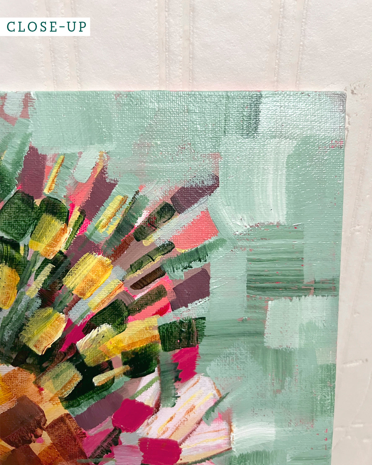 A close-up of an abstract floral painting by Jen Montgomery on canvas, showing textured brushstrokes in pink, red, yellow, green, and brown, leaning against a white wall on a light wood floor.
