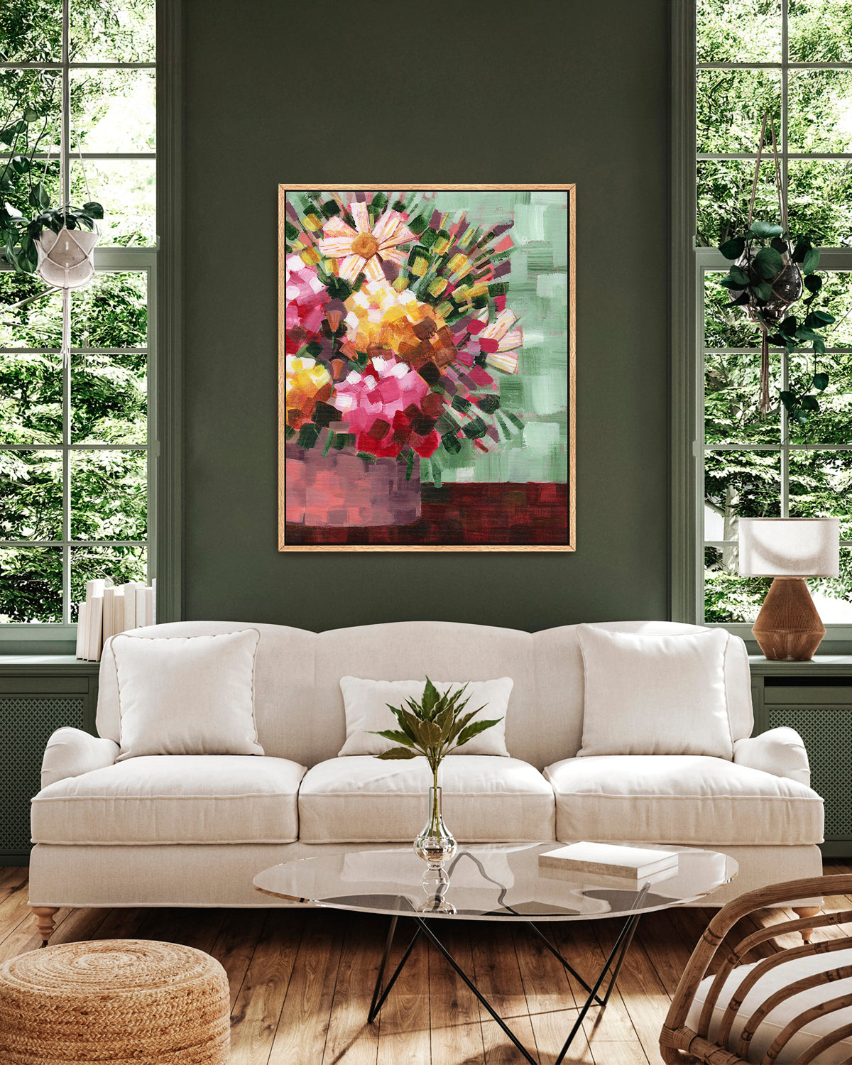 A framed abstract floral art print by Jen Montgomery featuring pink, yellow, red, and white flowers with green and brown accents, displayed above a sofa between two windows.