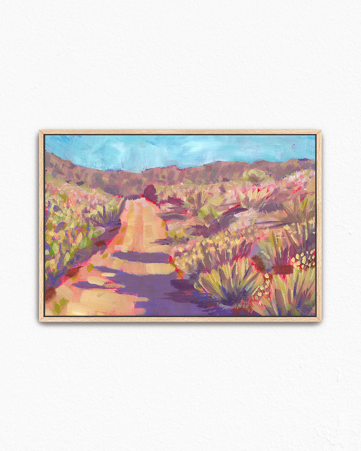 A framed landscape art print by Jen Montgomery of a desert hiking trail, featuring a sandy path winding through purple and green brush under a blue sky, displayed on a white wall.