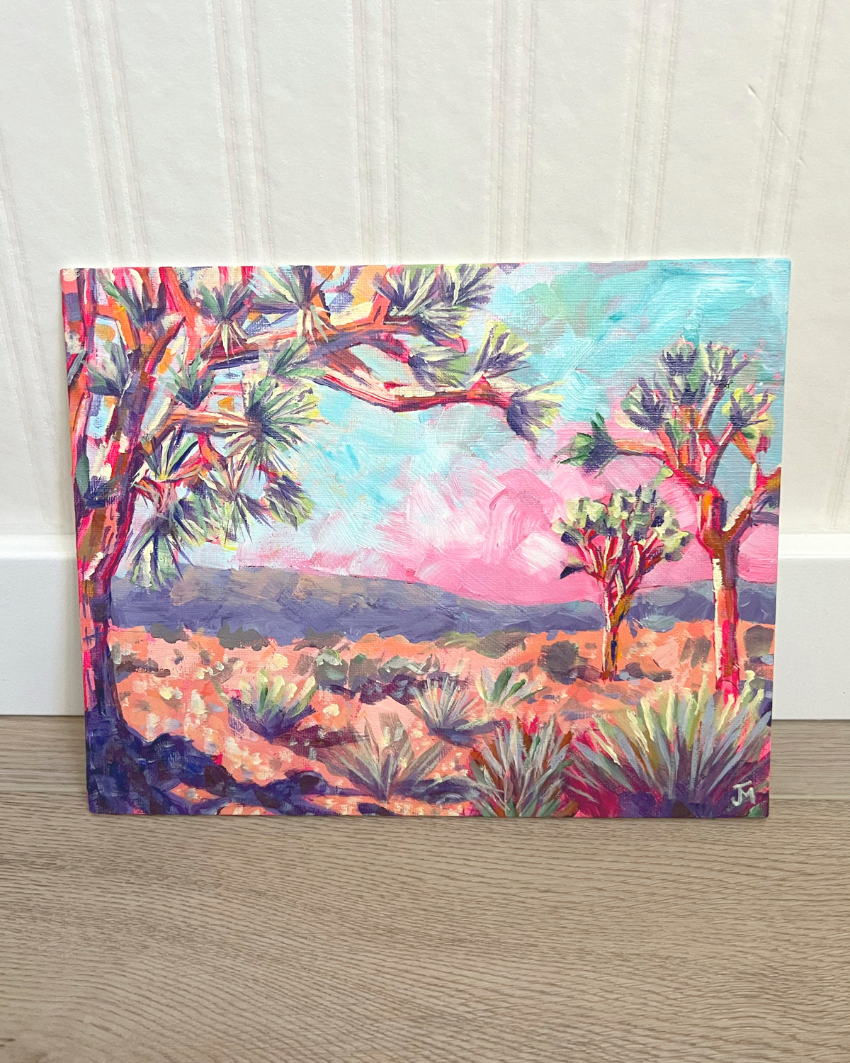 A painting by Jen Montgomery of a desert landscape with Joshua trees, painted in vibrant pink, blue, green, and orange tones, leaning against a white paneled wall on a light wood floor.