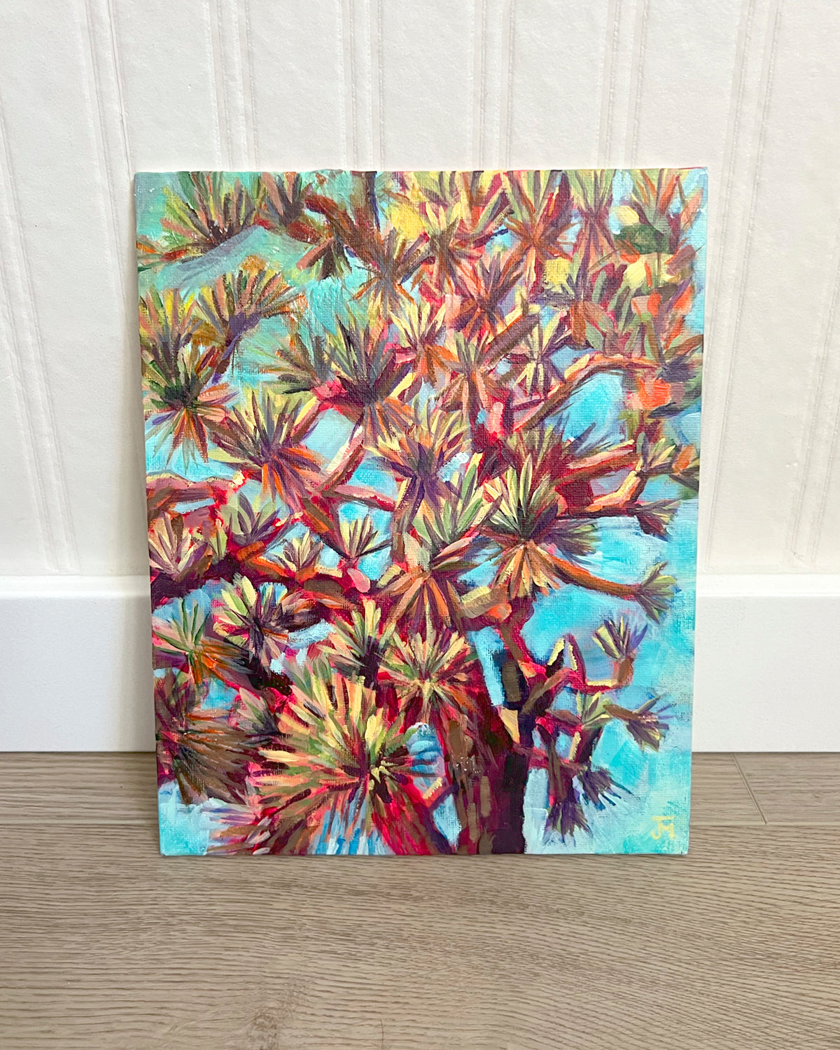 A vertical canvas painting by Jen Montgomery of a Joshua Tree with textured, starburst-like leaves in shades of orange, yellow, red, and green against a bright turquoise sky, displayed leaning against a white paneled wall on a light wood floor.