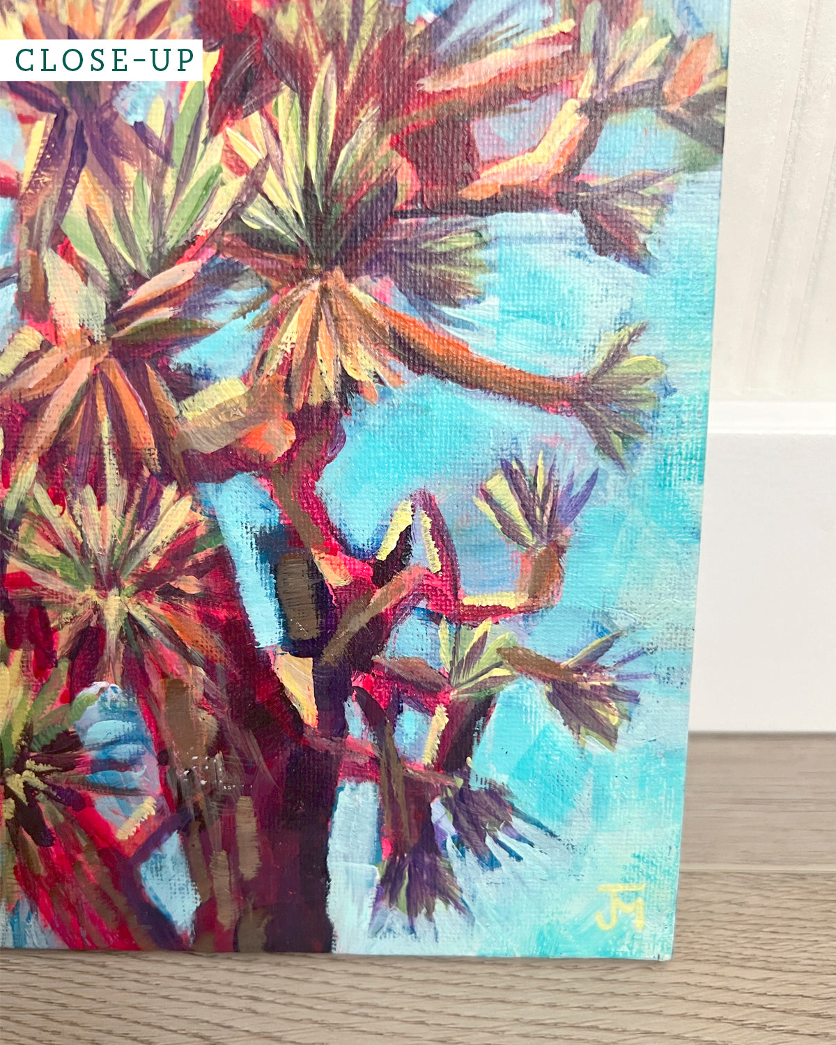 A close-up of a colorful painting by Jen Montgomery of a Joshua tree, showing textured brushstrokes in turquoise blue, red, orange, and green. A small yellow monogram signature is visible in the bottom right corner, and a 'CLOSE-UP' label is in the top left corner.