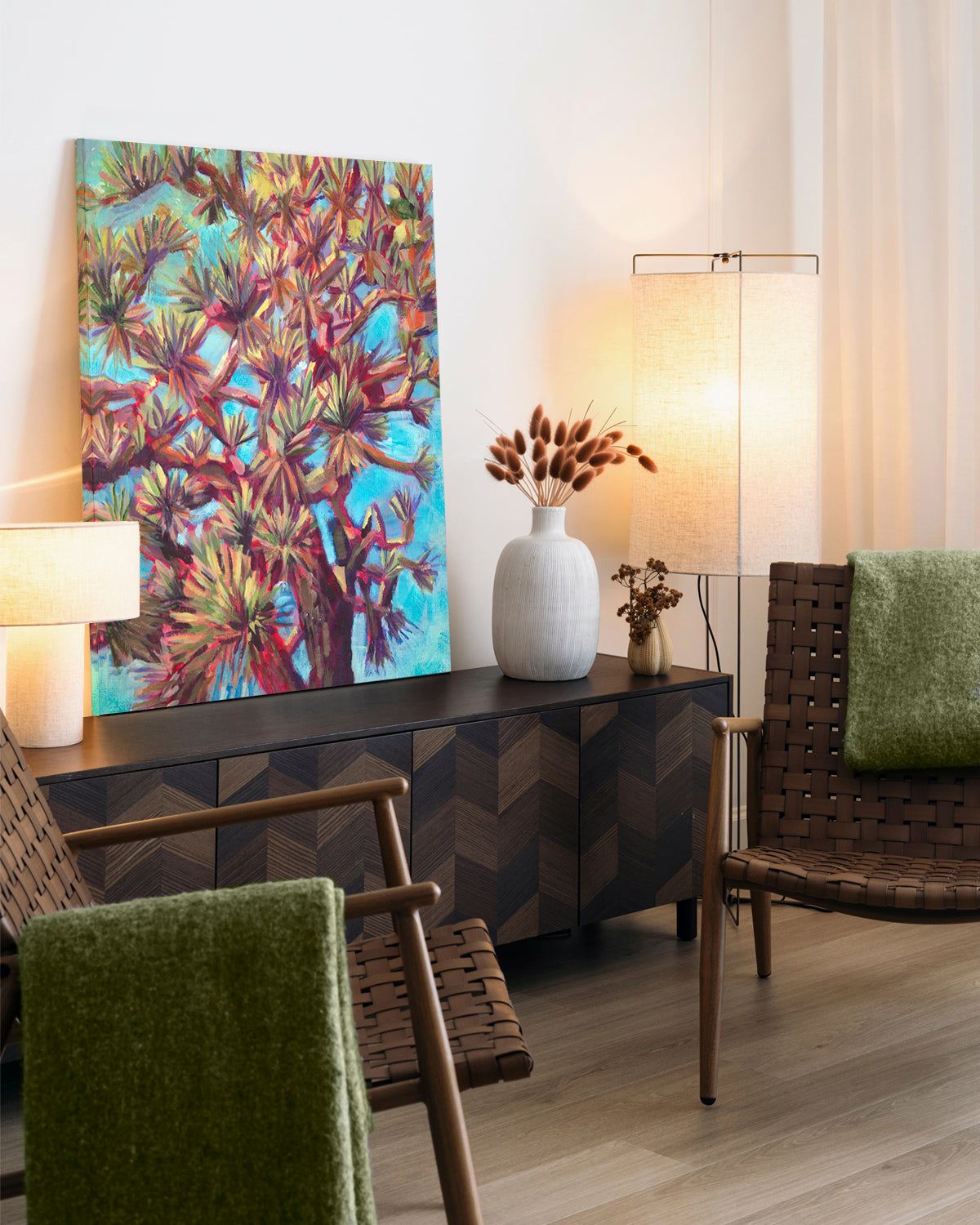 A colorful abstract canvas art print of Joshua Trees, displayed in a modern living room setting above a dark wood sideboard with lamps and chairs.