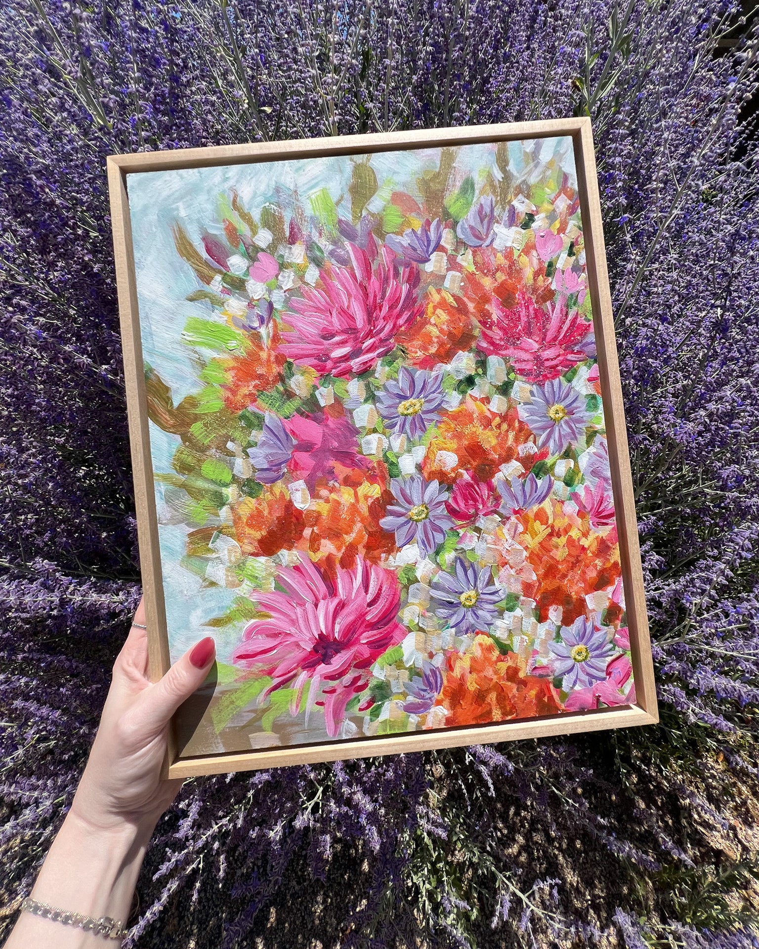 Jen Montgomery holds a framed painting of a colorful wildflower bouquet, set against a background of blooming lavender.