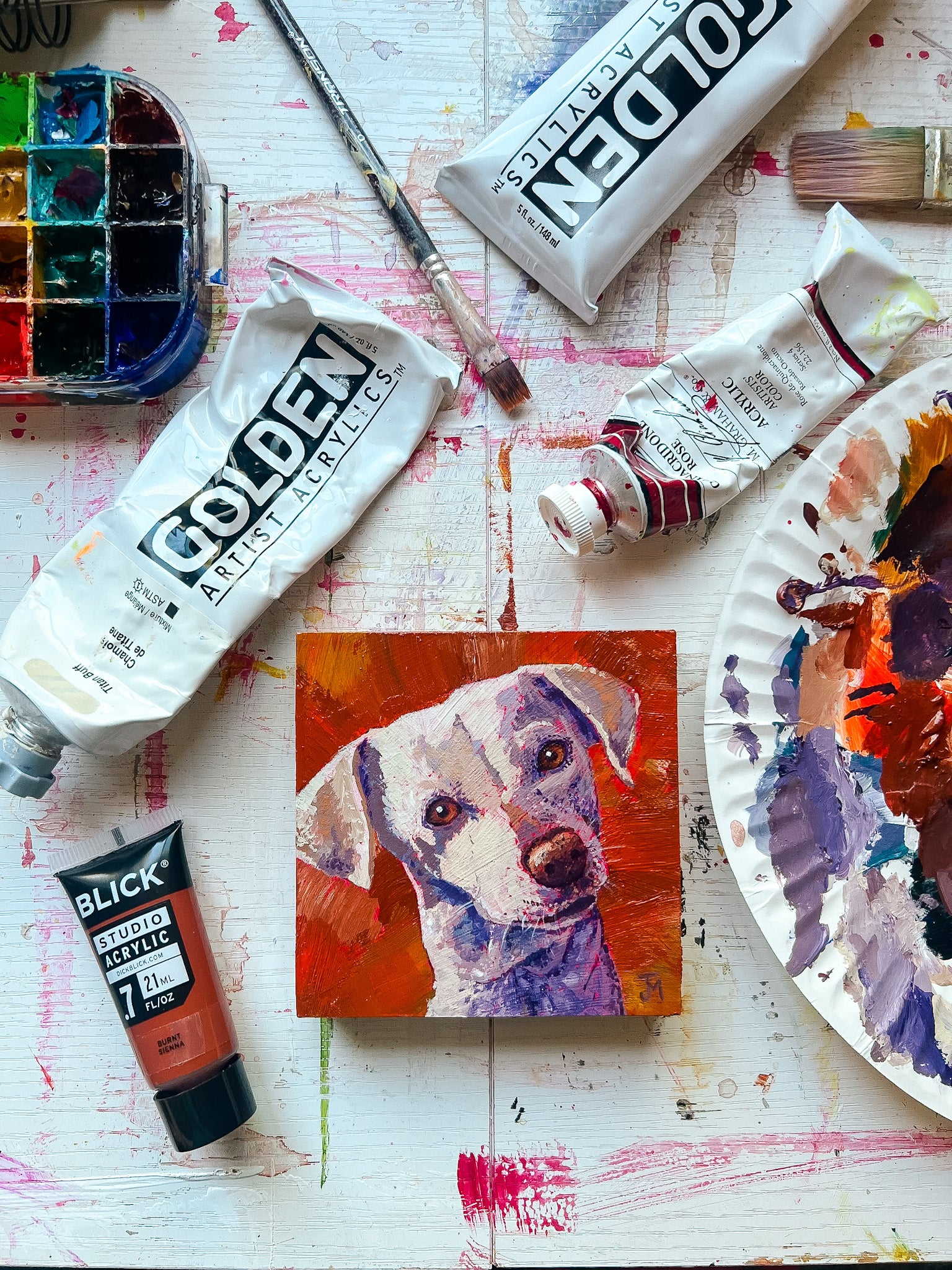 Artistic setup with paint tubes, a brush, and a small painting of a dog on a textured surface.