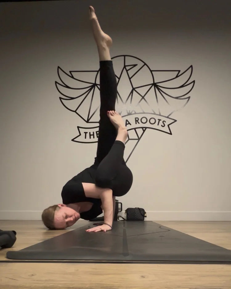 Jen Montgomery in fallen angel yoga pose on a black yoga mat in a studio, wearing a black short-sleeve top and black leggings. The background features a light gray wall with a black winged logo and the text 'THE YOGA ROOTS'.