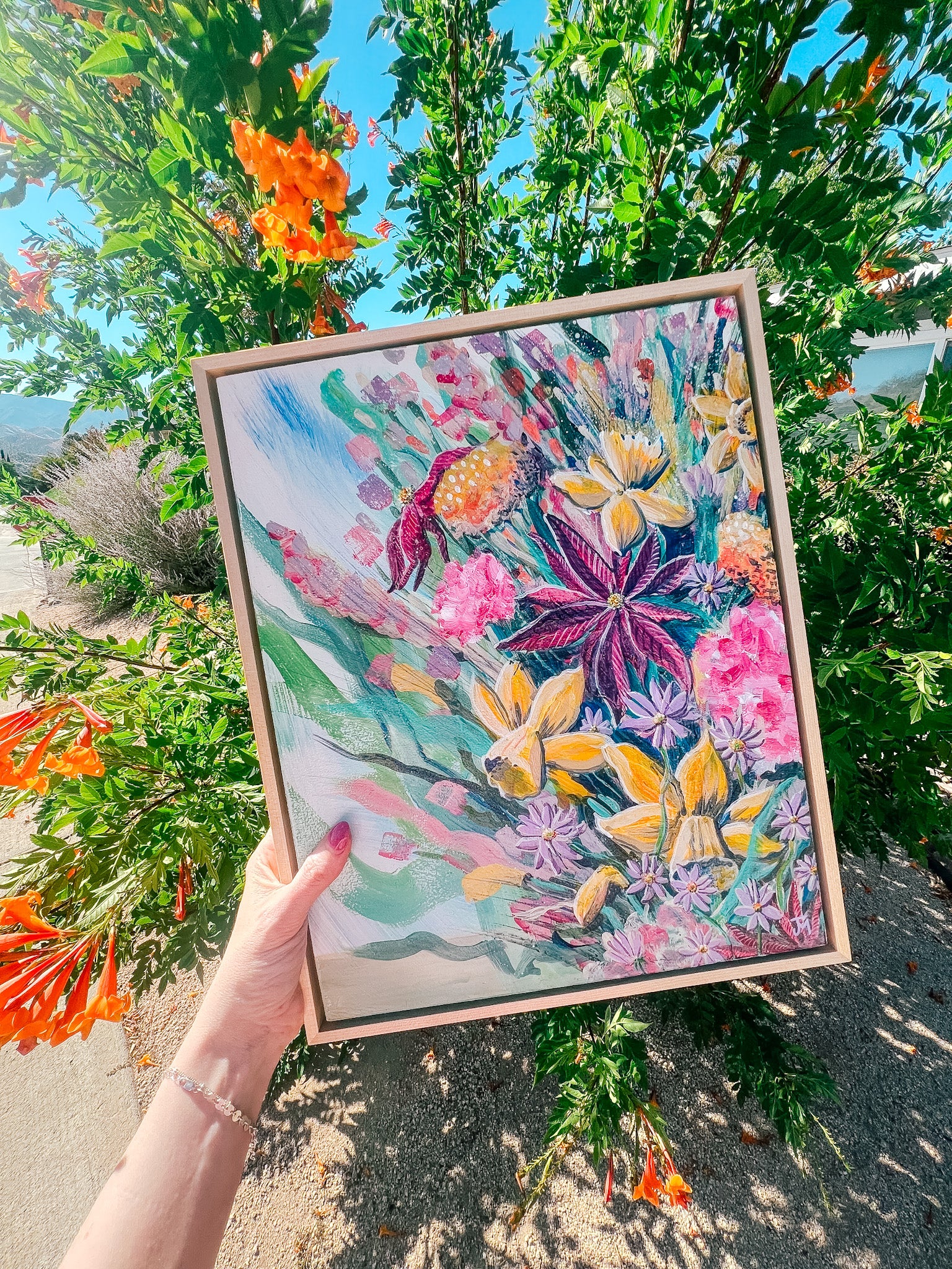 Framed floral borth month painting by Jen Montgomery held outdoors with greenery and flowers in the background