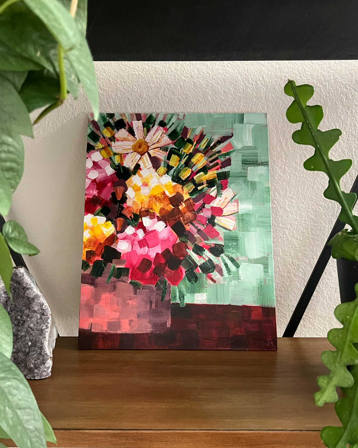 A colorful abstract floral canvas original painting by Jen Montgomery displayed on a wooden shelf, surrounded by green houseplants against a white wall.