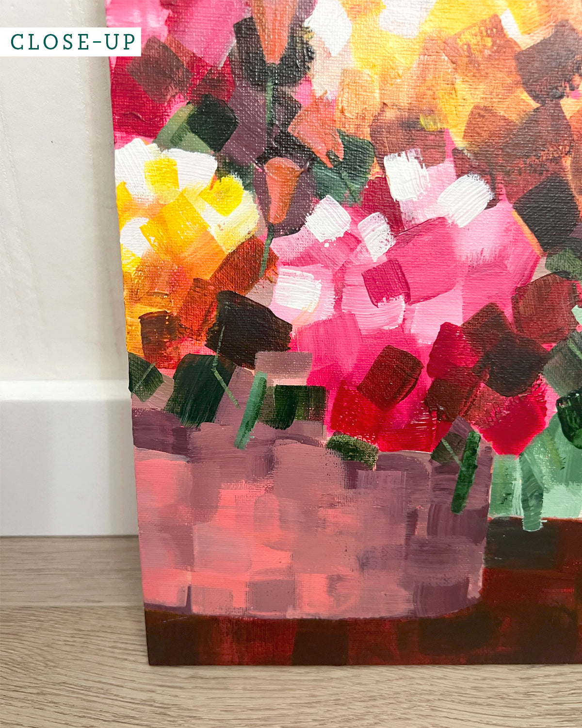 A close-up of an abstract floral painting by Jen Montgomery on canvas, showing textured brushstrokes in pink, red, yellow, green, and brown, leaning against a white wall on a light wood floor.