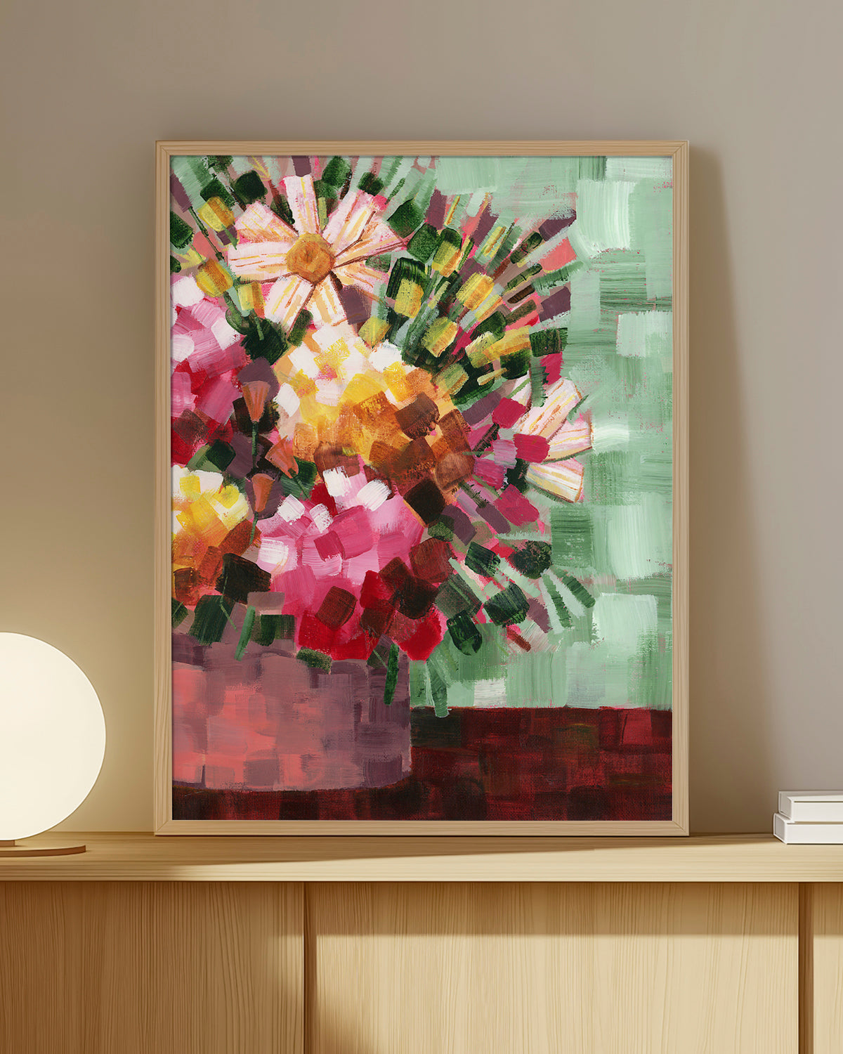 A framed abstract floral art print by Jen Montgomery featuring pink, yellow, red, and white flowers with green and brown accents, displayed on warm bookshelf.