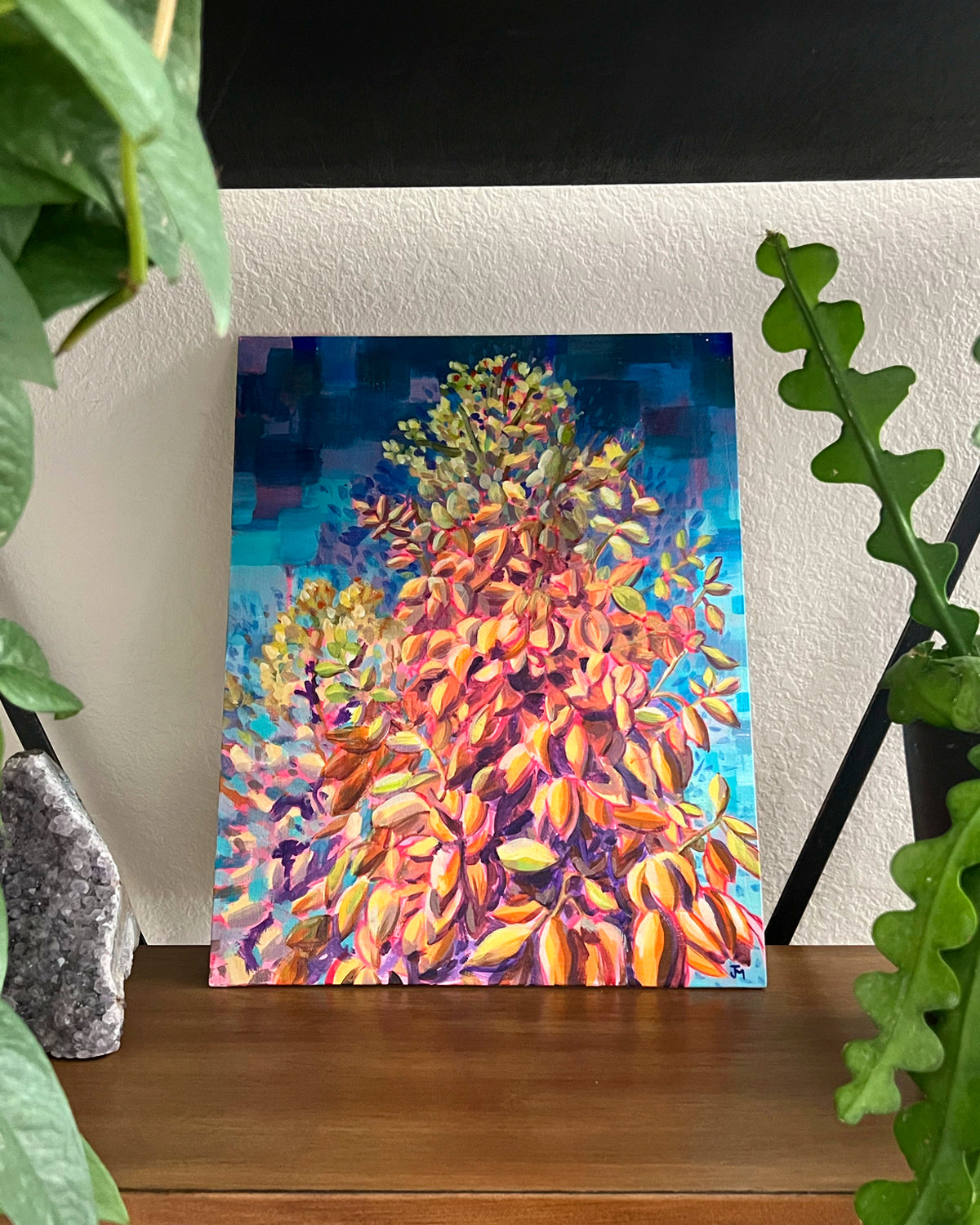 A colorful original painting by Jen Montgomery of a yucca plant with orange, yellow, and purple tones against a blue background, displayed on a wooden shelf between green houseplants.