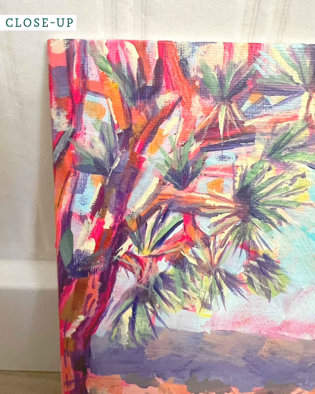 A close-up of a colorful painted desert landscape by Jen Montgomery on a textured canvas, featuring pink sky, two trees, and spiky desert plants. A small 'CLOSE-UP' label appears in the top-left corner.