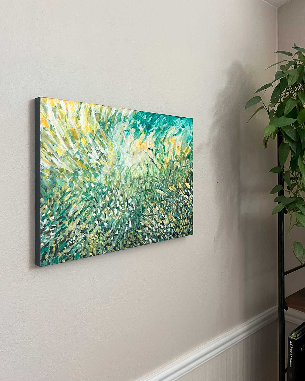 An abstract canvas painting by Jen Montgomery of green and yellow grasses or foliage, displayed on a beige wall in a home interior next to a green houseplant.