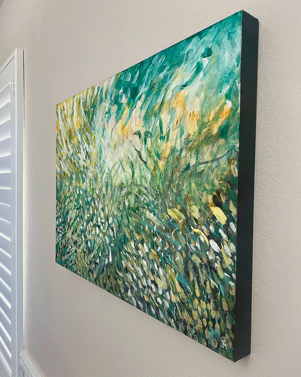 An abstract landscape canvas painting by Jen Momtgomery featuring textured brushstrokes of green, teal, yellow, and white, displayed on a light beige wall next to a white shutter.